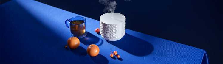 An aroma nook essential oil diffuser placed on a blue tablecloth next to a cup of tea and two clementines