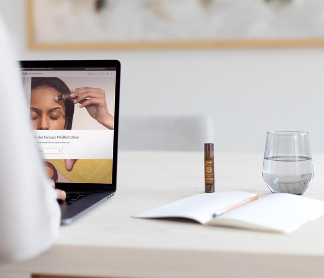 A person works on a laptop showing a Saje roll-on with an open notebook, roll-on, and glass of water by their side. 