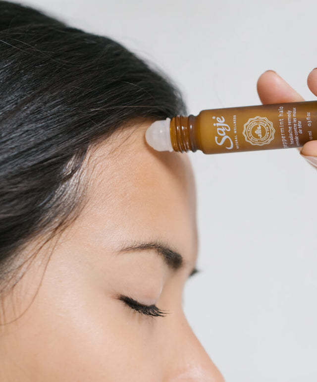 A person with dark hair and closed eyes applies a roll-on to their hairline against a white wall backdrop.