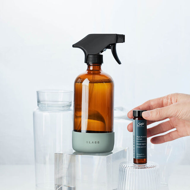 A hand removing the cap of the refill, next to the glass spray bottle on a glass pedastal