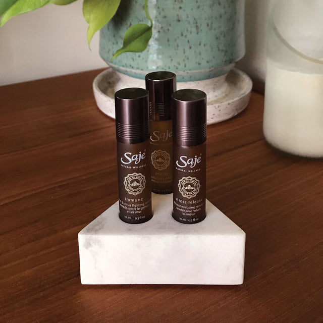 Three roll-ons inside the triangle marble holder on a wooden desk with a sea-foam speckled vase with greenery behind it