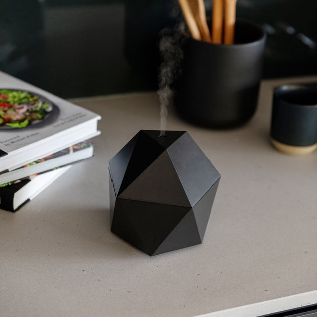 The black diffuser on a desktop surrounded by cookbooks and some pencils in a cup holder.