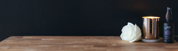 Saje body mist, a rose-gold candle, and a white rose sitting on a desk