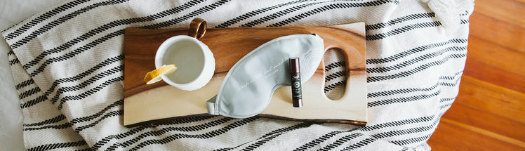 A Saje roll-on with an eye mask, on a wooden board on top of a stripped sheet