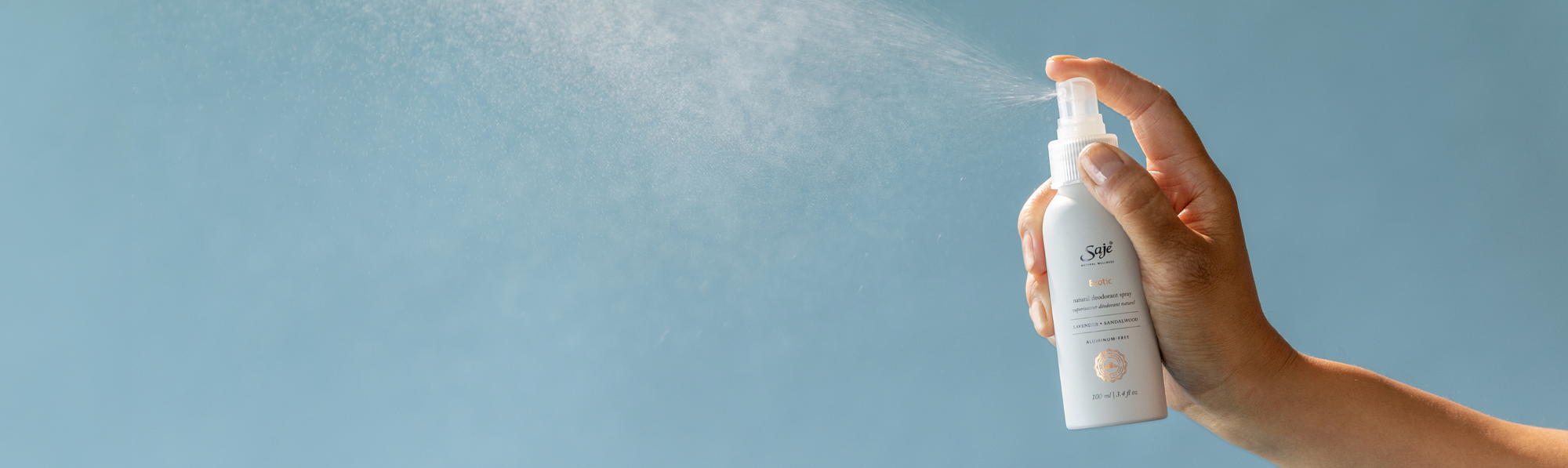 A hand spraying a bottle of Exotic Natural Deodorant Spray against a blue background