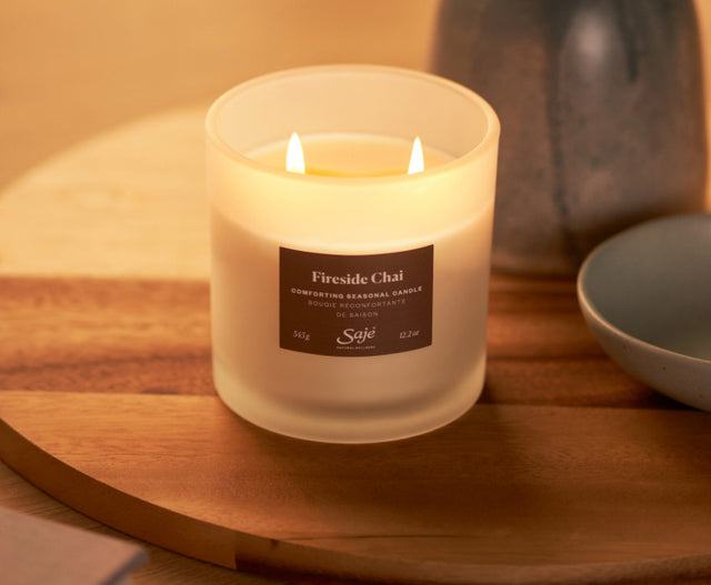 A lit double-wick candle with a brown label sits on a side table beside a couch and mug in a cozy, dim room.