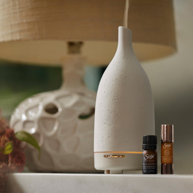 Aroma Om Diffuser in white, Liquid Sunshine diffuser blend and Peppermint Halo Roll-On sitting on a white surface next to a lamp.