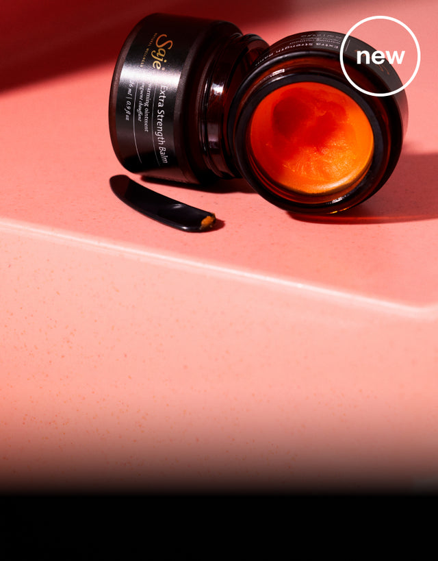 Two open jars of Saje Extra Strength Balm show off the striking bright orange balm against a dark pink background. 