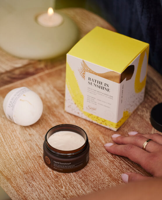 Liquid Sunshine body butter and bath swish are styled on a wood table beside a decorative holiday box.
