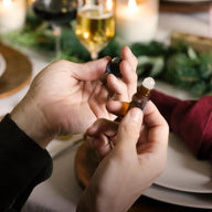 A man, sitting at a stylized dinner table, unscrews the cap off a Saje roll-on.