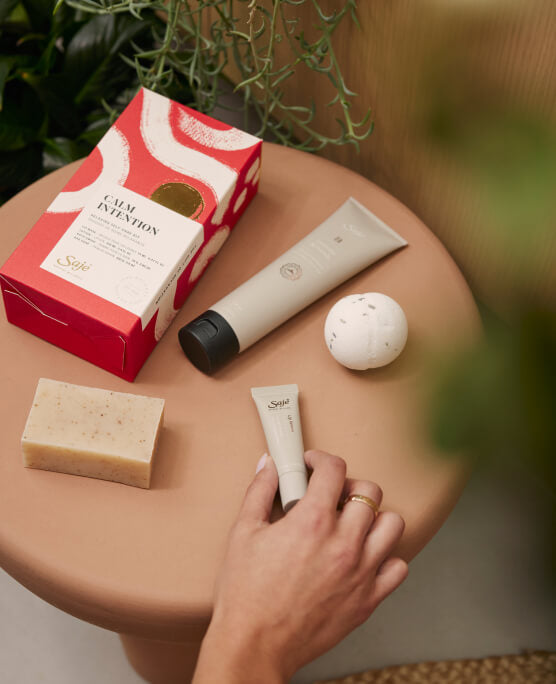 Calm Intention Kit, with a lotion, lip mask, bath swish, bar soap, and decorative box, is arranged flat on a wood table.