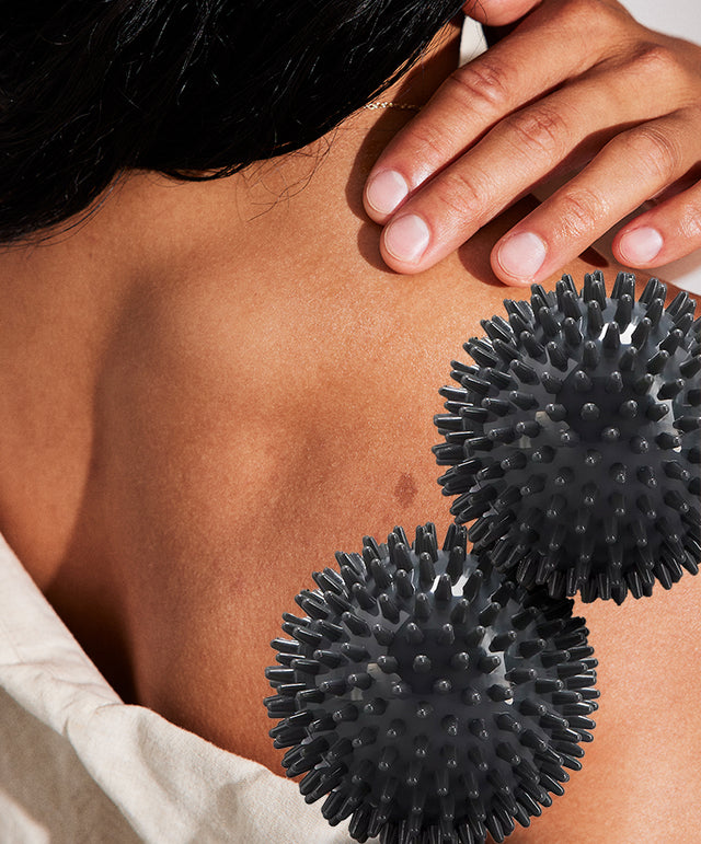 Close-up of a person’s fingers massaging their back; two dark grey, spiky massage balls are shown beside. 