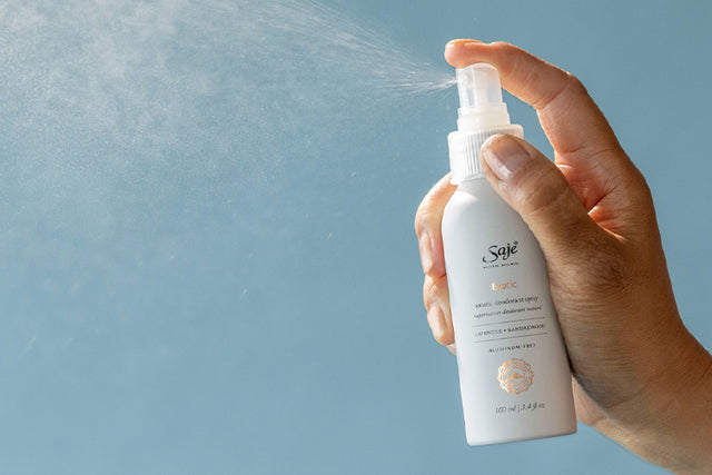 A hand sprays Saje exotic natural deodorant in front of a blue background