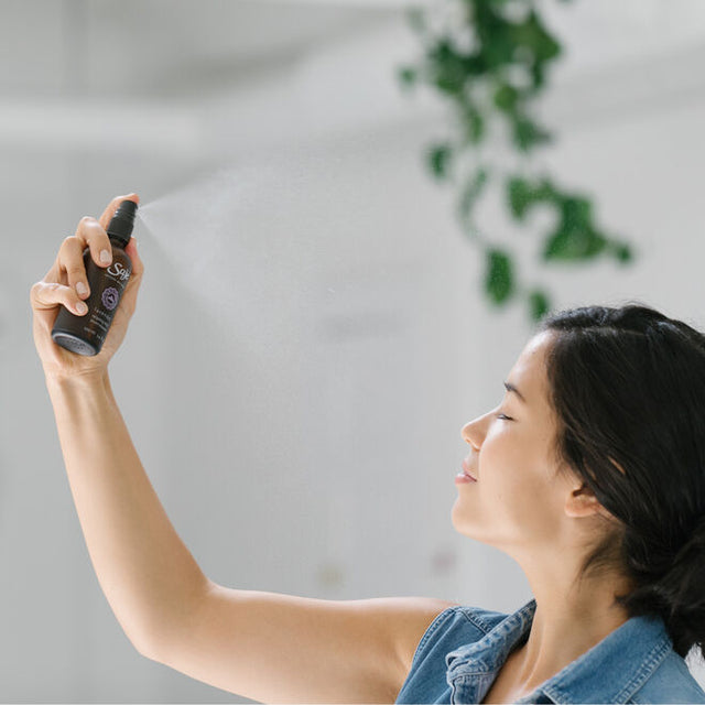 a woman in a denim sleeveless shirt holding a mist bottle and spraying it towards her face with her eyes closed