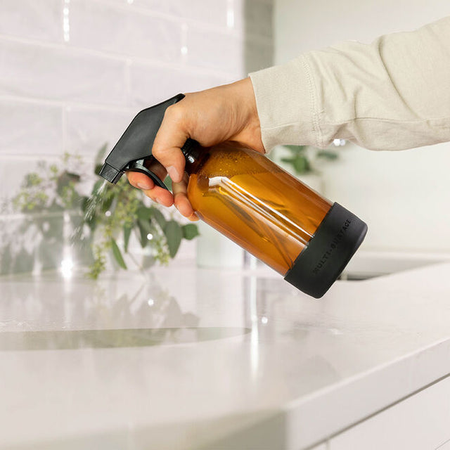 a hand spraying the multi-surface cleaning solution from the glass spray bottle onto a white countertop