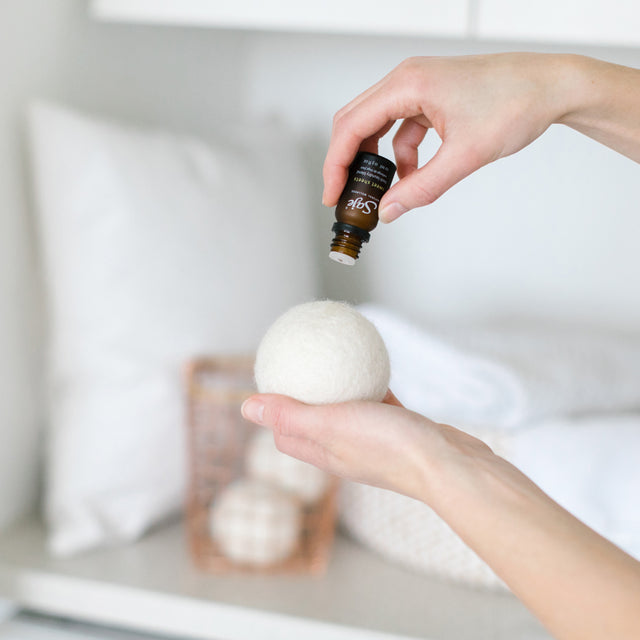 A pair of hands, one holding a dryer ball and another with the uncapped Sweet Sheets laundry blend, putting essential oil drops onto the dryer ball