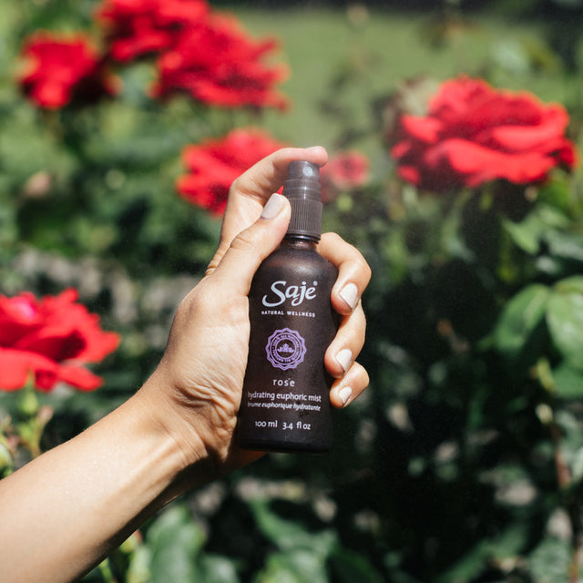 A person holding Rose Hydrating Euphoric Mist 100ml in front of a garden of flowers 