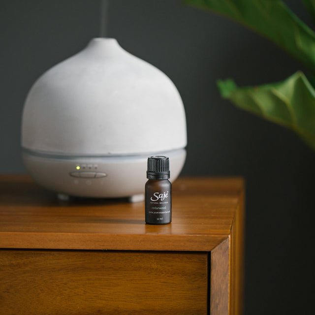 Cedarwood single note essential oil brown bottle on a wooden cabinet with an Aroma Om Deluxe white diffuser behind on a grey background 