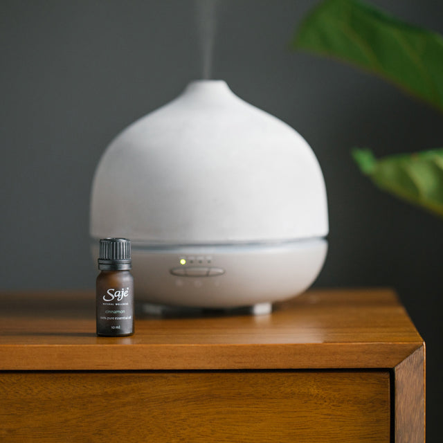 Cinnamon single note essential oil brown bottle with the cap on on a wooden side table in front of a white Aroma Om Deluxe. The background colour is a dark charcoal