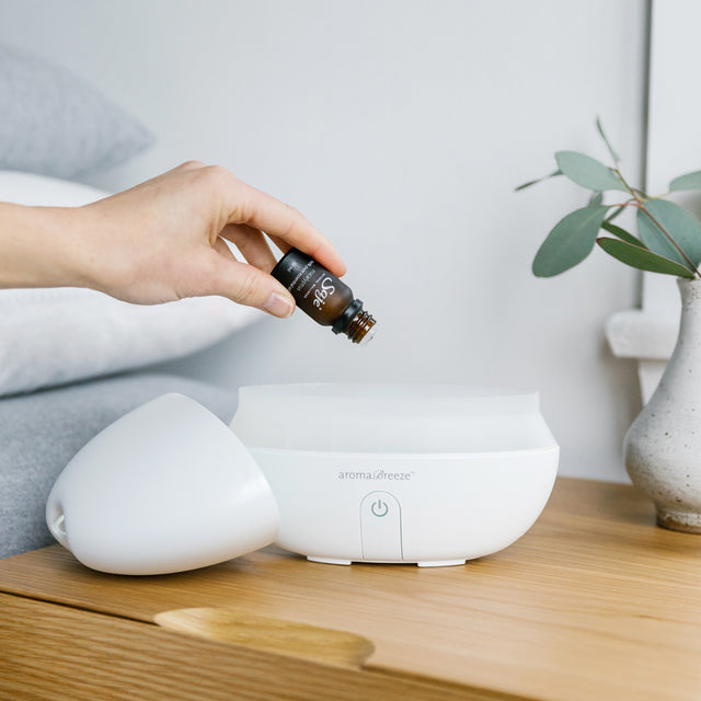 a hand holding an uncapped bottle over a diffuser water tank on a bedside table